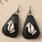 Black  leather teardrop-shaped earrings with cowrie shells  in the middle. The cowrie shell is surrounded with black and white beads on a beige fabric background