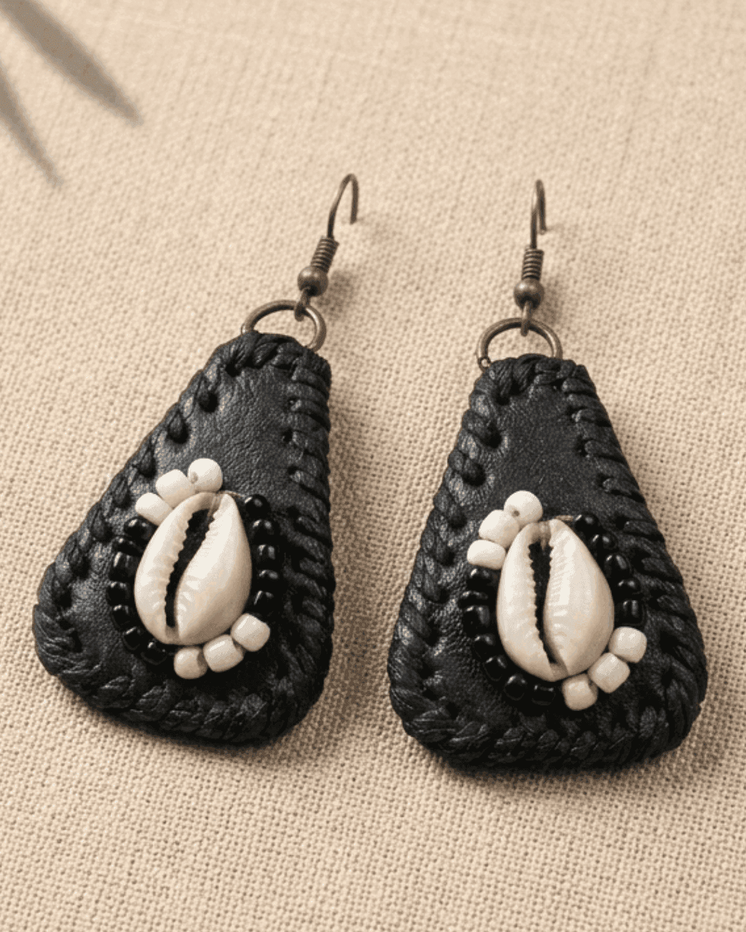 Black  leather teardrop-shaped earrings with cowrie shells  in the middle. The cowrie shell is surrounded with black and white beads on a beige fabric background