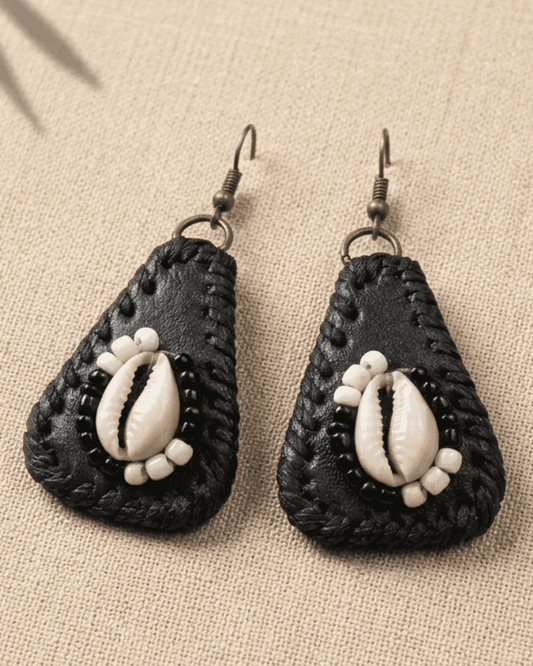 Black  leather teardrop-shaped earrings with cowrie shells  in the middle. The cowrie shell is surrounded with black and white beads on a beige fabric background